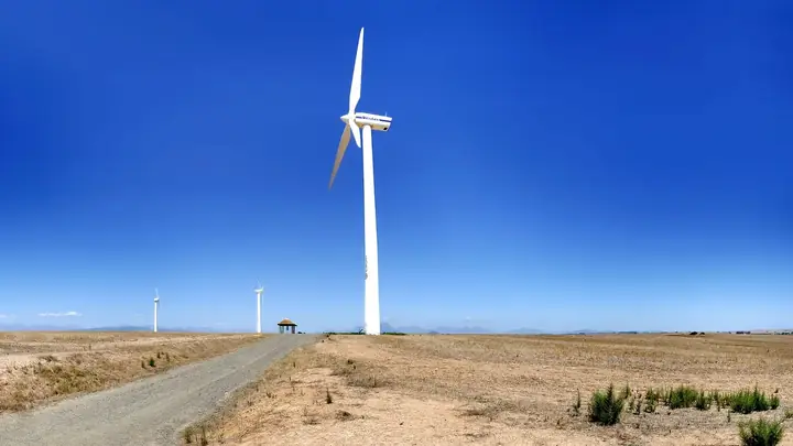 A white wind turbine with the Eskom logo on its tower stands beside a gravel road on dry farmland, with two smaller turbines in the distance